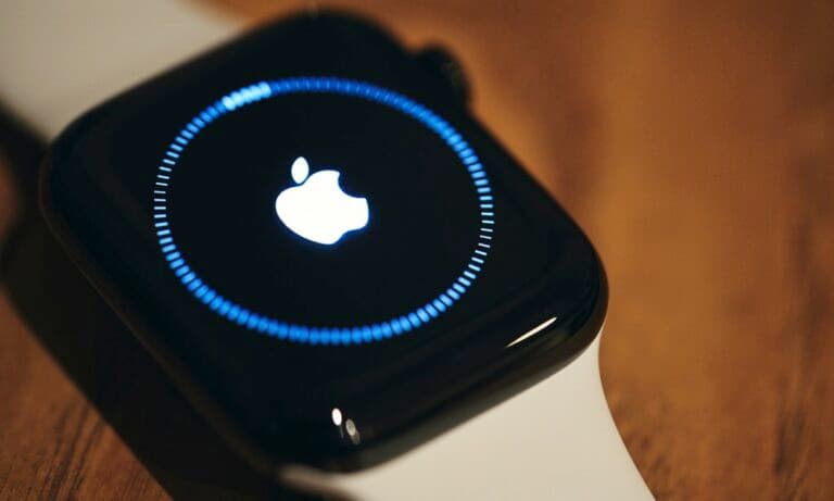Close-up of an Apple Watch on a wooden surface with the Apple logo centered on its screen. A blue circular progress ring surrounds the logo, indicating an update process to the watchOS 11.3 beta 2 version. The watch has a white sport band and a glossy black case